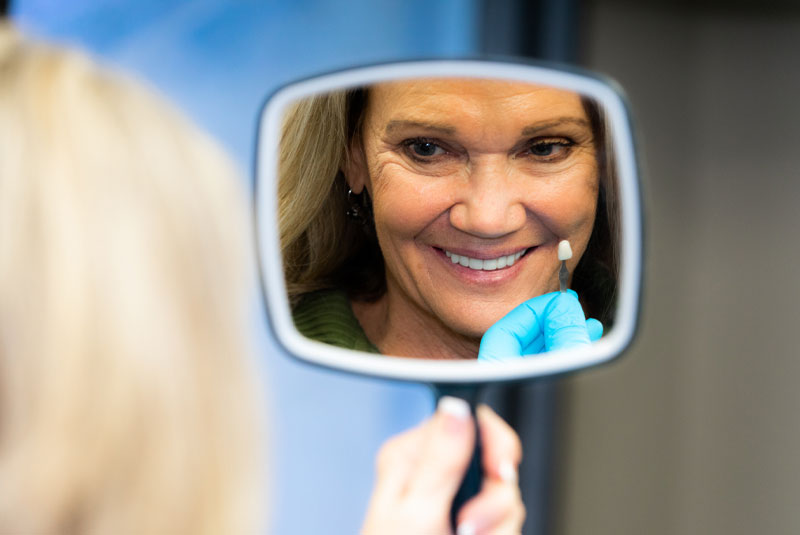 Jennifer, All on 4 Dental Implants Patient, Smiling Into A Mirror With A Sample Tooth